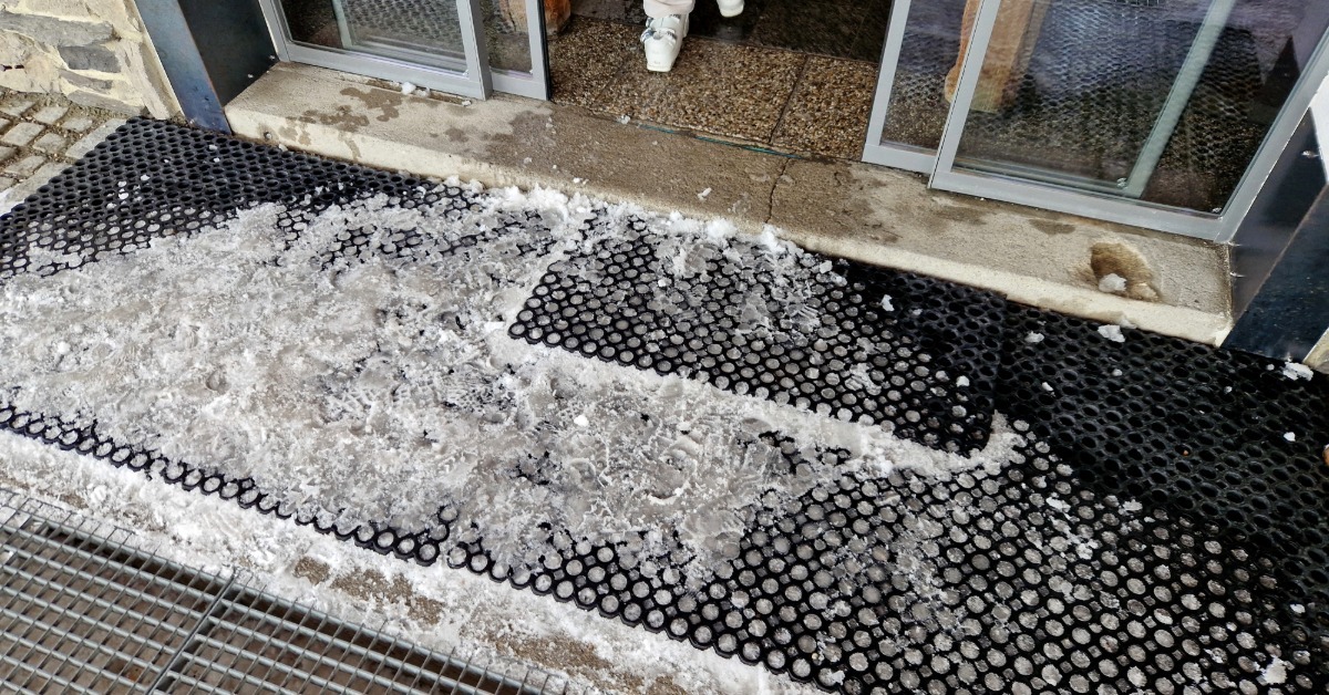 A black honeycomb rubber entrance mat lying across a floor near sliding doors, with snow and debris in the holes.