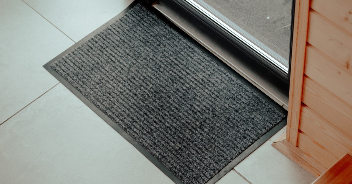 A textured black doormat sitting near a building entrance on a light floor, with sunlight falling across the surface.