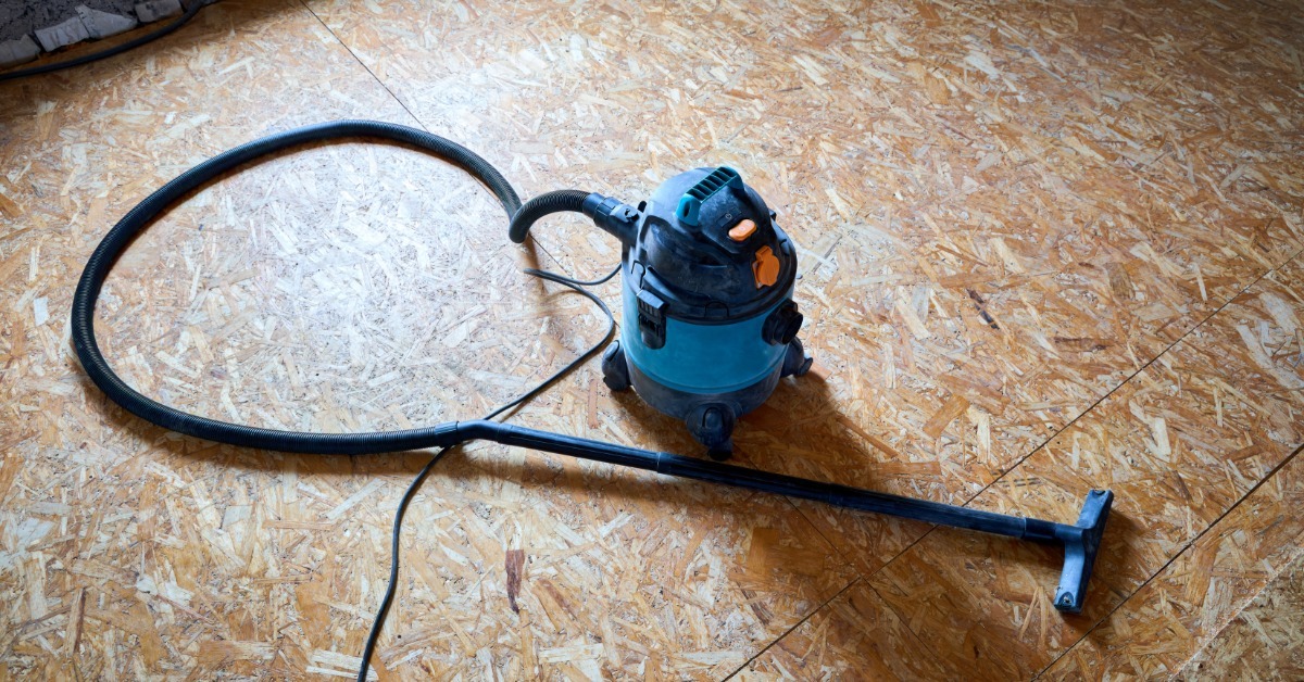 A commercial-grade shop vac sits on a wooden, unfinished floor board. The long suction hose lays down.