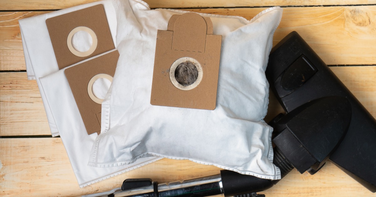 A vacuum head on the floor next to a full vacuum bag. There are two unused vacuum bags underneath the full bag.