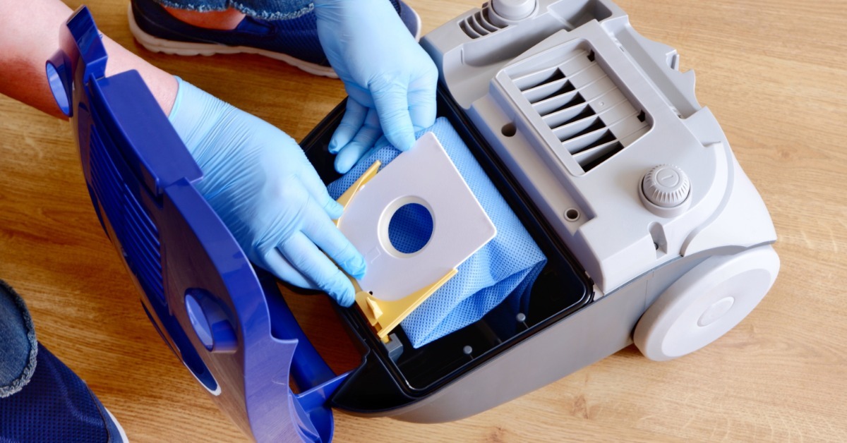 Someone wears blue latex gloves as they replace the bag in a vacuum cleaner. They wear jeans and sneakers.
