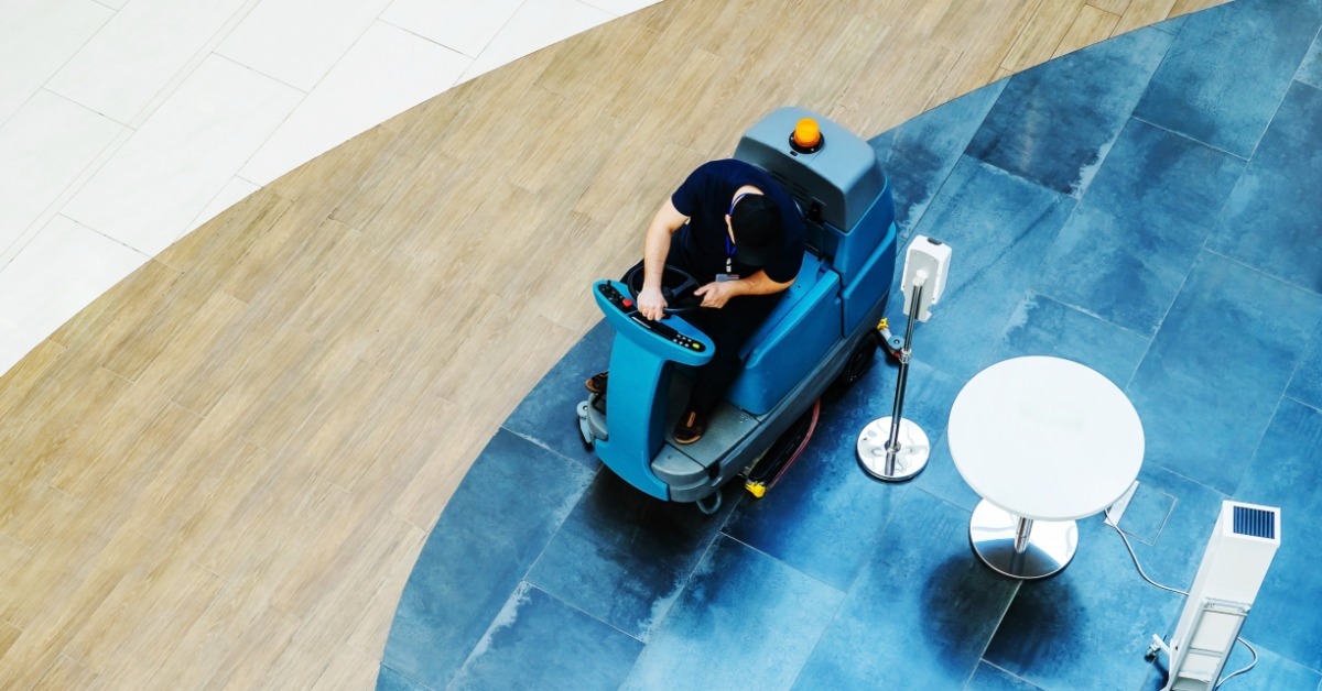 A commercial space with a professional cleaning the floors with a ride-on vacuum. There's a cafe table next to him.