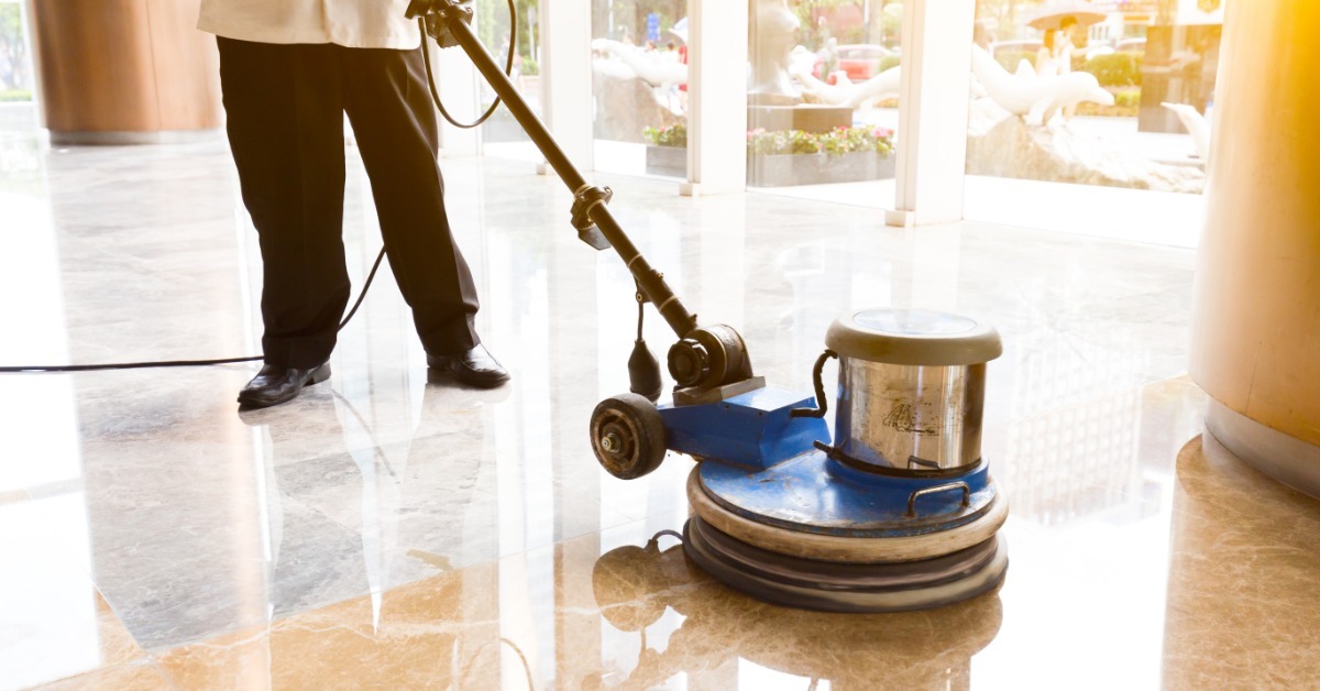 A professional floor cleaner uses a floor polishing machine in a commercial space. They wear black pants.