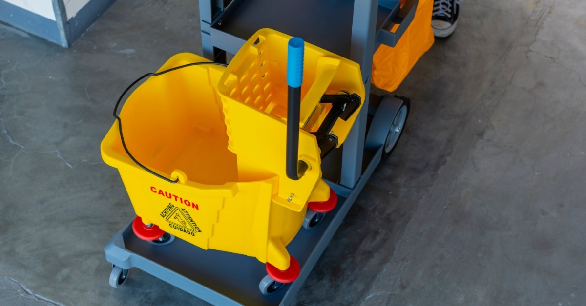 An industrial yellow mop bucket with nothing in it. The bucket sits on a janitorial cart in a business hallway.