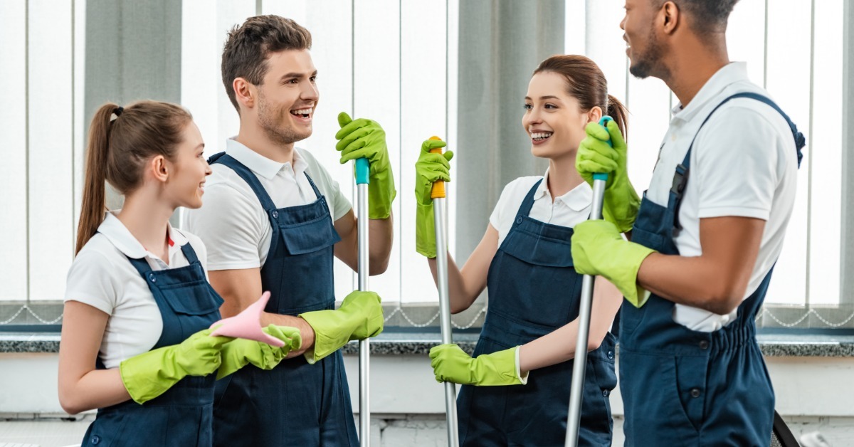 Four cleaners wearing gloves and uniforms standing together indoors while holding mop handles and talking.