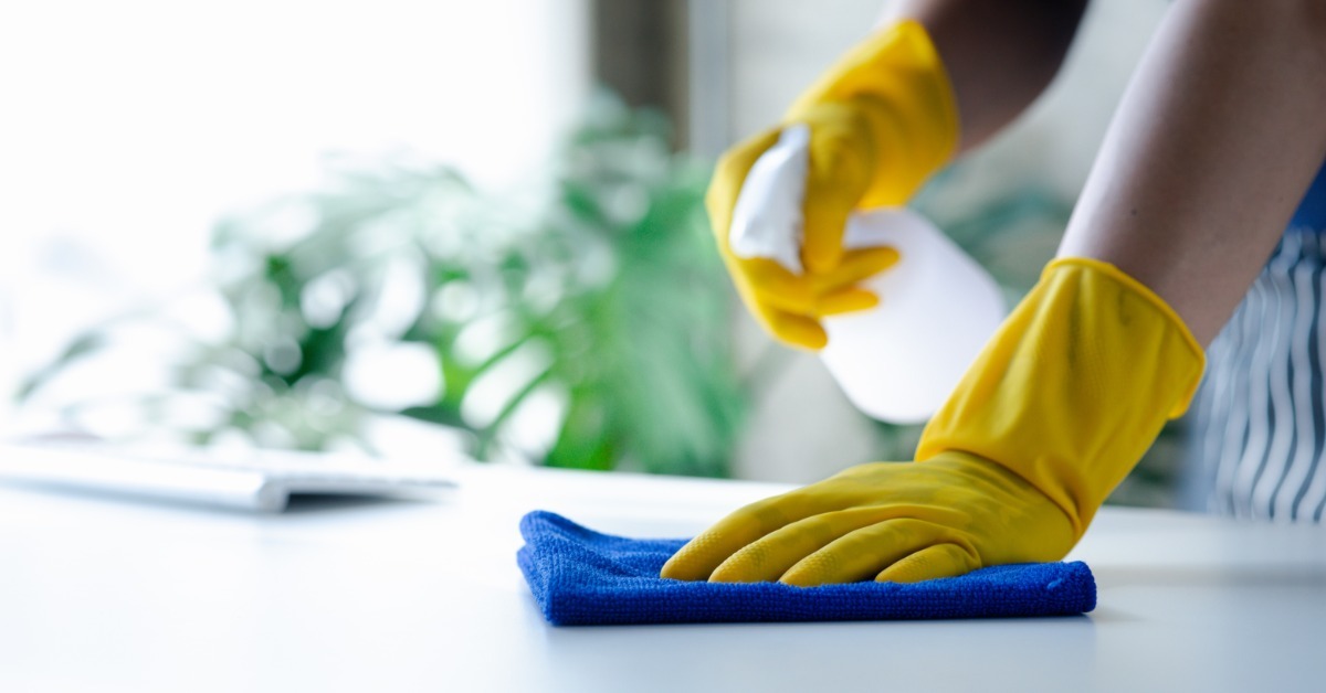 A person wearing yellow gloves cleaning a white tabletop with a blue cloth and holding a spray bottle.