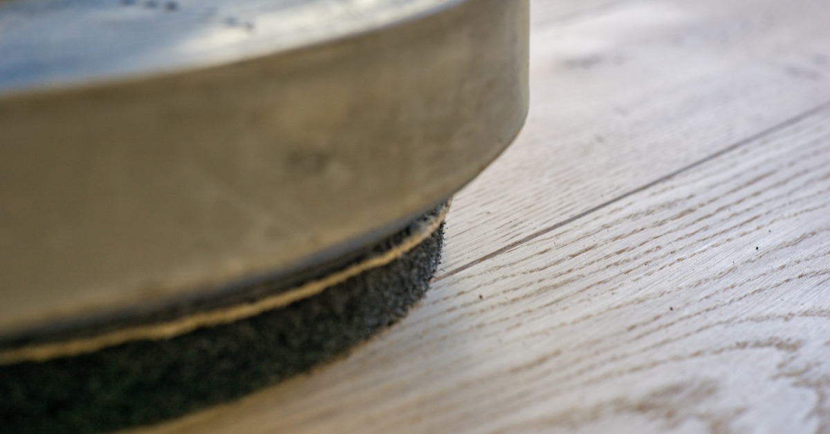 A close-up of a floor buffer machine on the floor in a room. The machine is silver with a black pad attached.