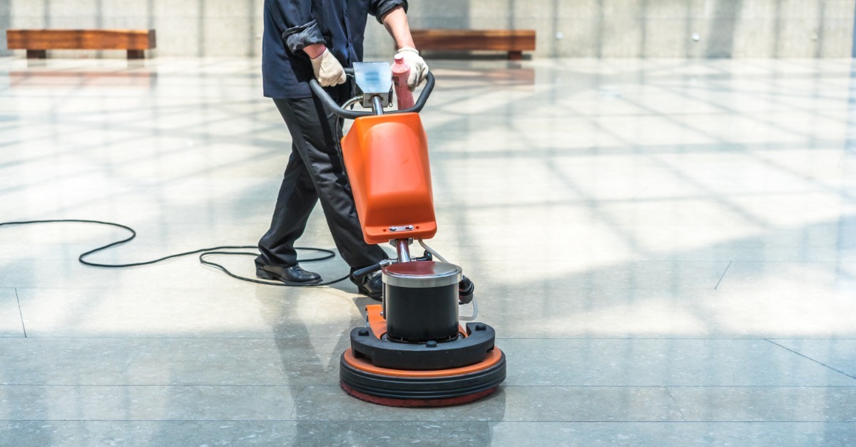 A commercial maintenance professional cleans the floors with a red floor buffing machine. They wear a uniform.