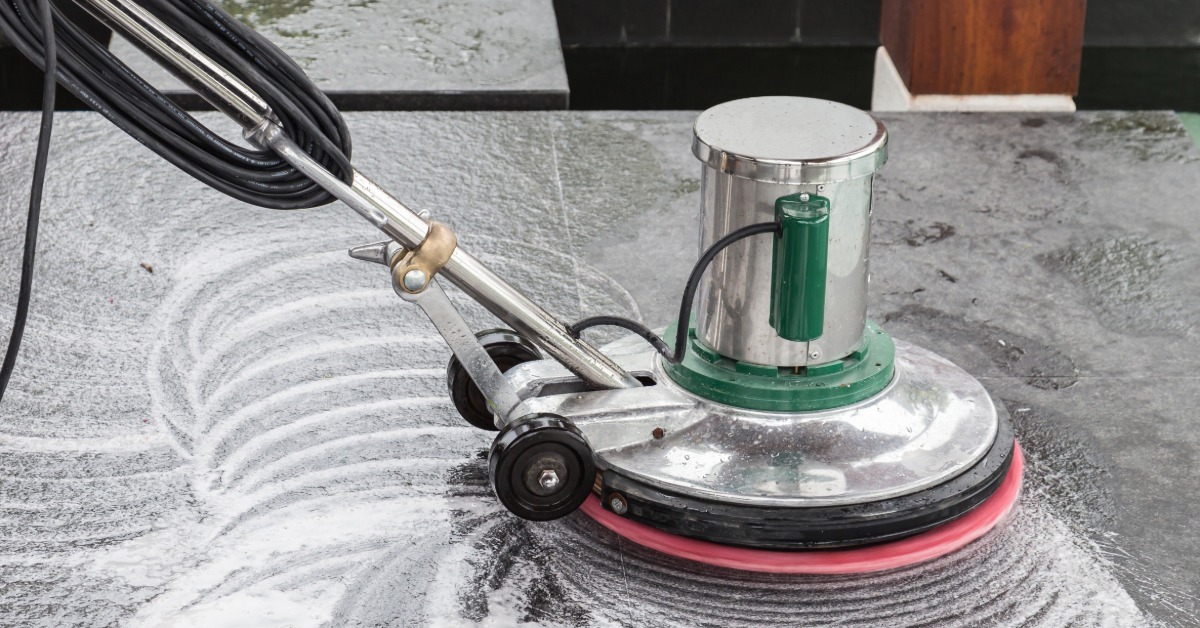 A close-up of a floor cleaning machine scrubbing the ground underneath it. There are suds and chemicals on the floor.
