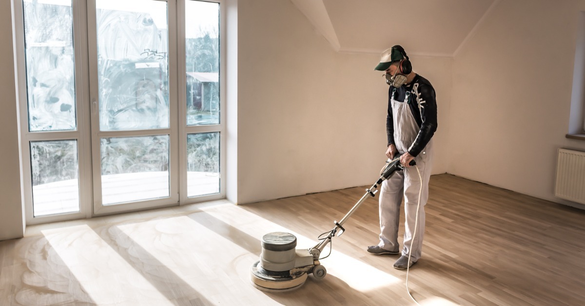 A flooring professional wears PPE and face protection as they take a flooring machine across a residential floor.