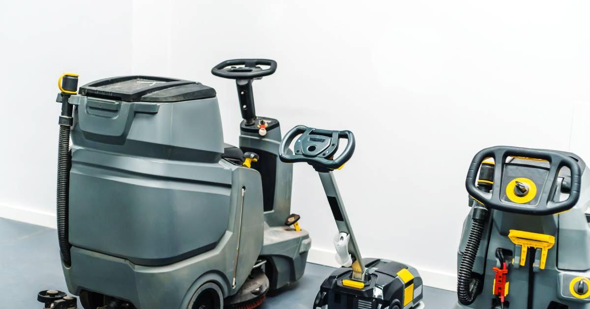 Three different types of floor cleaning machines lined up next to each other. They are gray with black and yellow trim.
