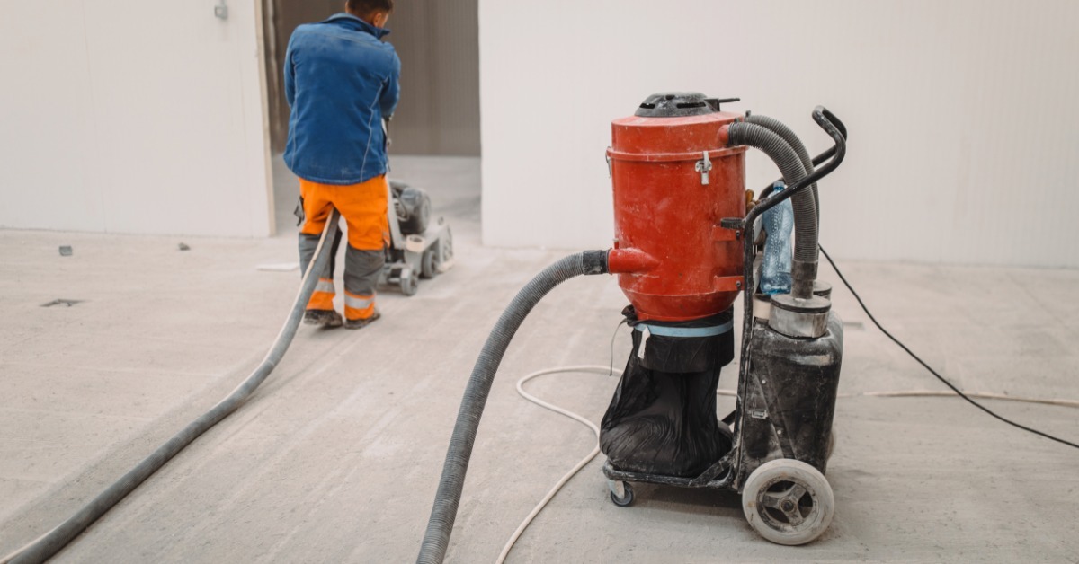 A professional flooring person wears a blue jacket and orange pants as they run a concrete polisher across the floor.